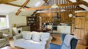 A spacious living area with a sofa, armchair, and coffee table. A kitchen with wooden cabinets and a dining table is visible in the background. - Beautiful Barn Conversion With Wood-burning Stove (Yeovil)