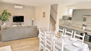 Una sala de estar moderna con un sofá gris, un televisor de pantalla plana y un comedor para cuatro con sillas blancas y una mesa.