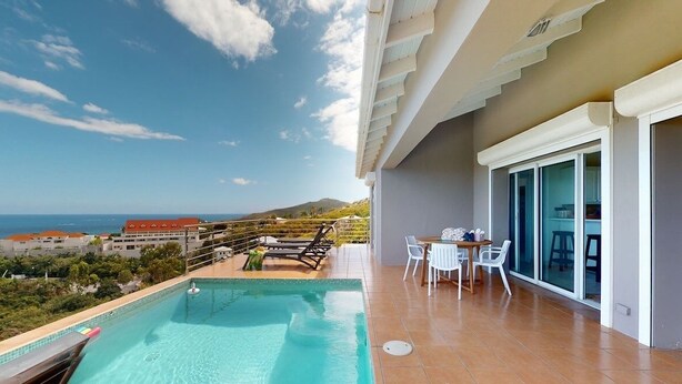 A balcony with a pool, a table with chairs, and a view of the ocean and buildings. - ☀️🌴Breezy Bird by IRE Vacations - Cozy 2 bedroom Villa with Private Pool (Oyster Pond)