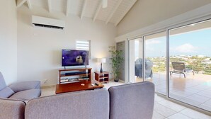 A living room with a flat-screen TV, grey sofas, a wooden entertainment unit, and a balcony with a view of the city. - ☀️🌴Breezy Bird by IRE Vacations - Cozy 2 bedroom Villa with Private Pool (Oyster Pond)