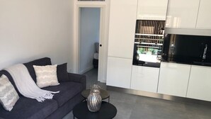Ett modernt vardagsrum med en mörkgrå soffa, ett litet runt bord och en dekorativ vas. Intill vardagsrummet finns ett kök med vita skåp och en svart bänkskiva.