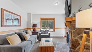 A living room with a grey sofa, a wooden coffee table, a grey armchair, a stone fireplace, and a large window with blinds. - Condo in River Run Village Only Walking Distance to Surrounding Lifts! (Keystone)
