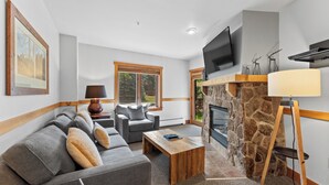 A modern living room with a stone fireplace, grey sofas, a coffee table, and a flat-screen TV mounted above the fireplace. - Condo in River Run Village Only Walking Distance to Surrounding Lifts! (Keystone)
