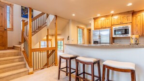 A kitchen with wooden cabinets, a stainless steel refrigerator, a microwave, and a granite countertop. There are two wooden stools with cushioned seats.