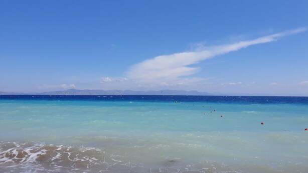 Una playa con agua turquesa cristalina, una orilla arenosa y montañas distantes bajo un cielo azul.