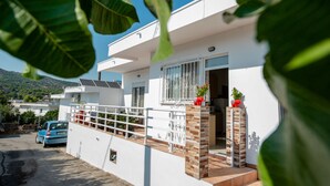 Una casa moderna de dos pisos con fachada blanca, un balcón con barandillas y un pequeño jardín con plantas en macetas.