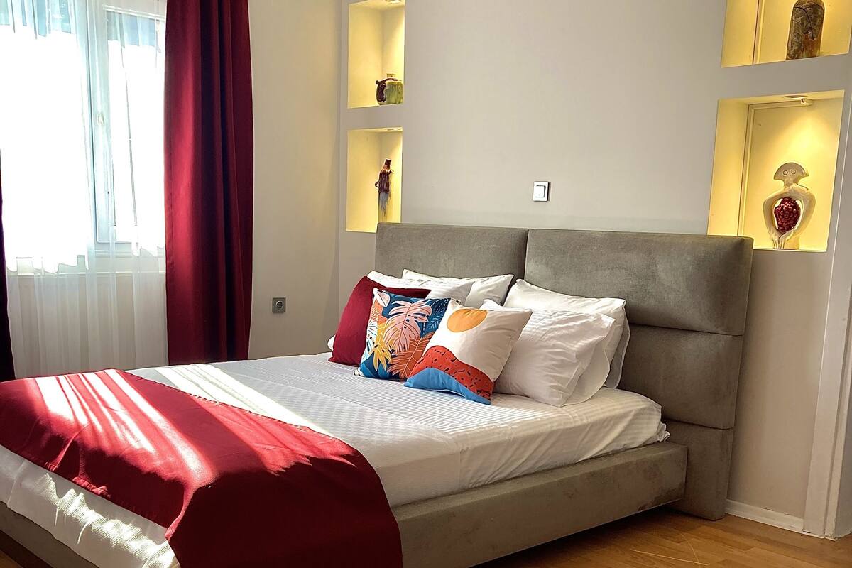 A modern bedroom with a large bed, wooden flooring, and a view of the city through the window.