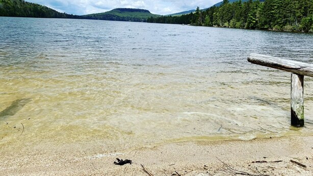 A clear lake with a wooden bench on the shore, surrounded by forested hills. - Snowmobile & ATV right on our property to trails, Ride the wilds, corridor 5. (Stark)