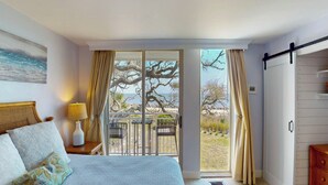 Un dormitorio con cama, mesita de noche, lámpara, vistas al exterior a través de una puerta corrediza de vidrio y cómoda de madera.