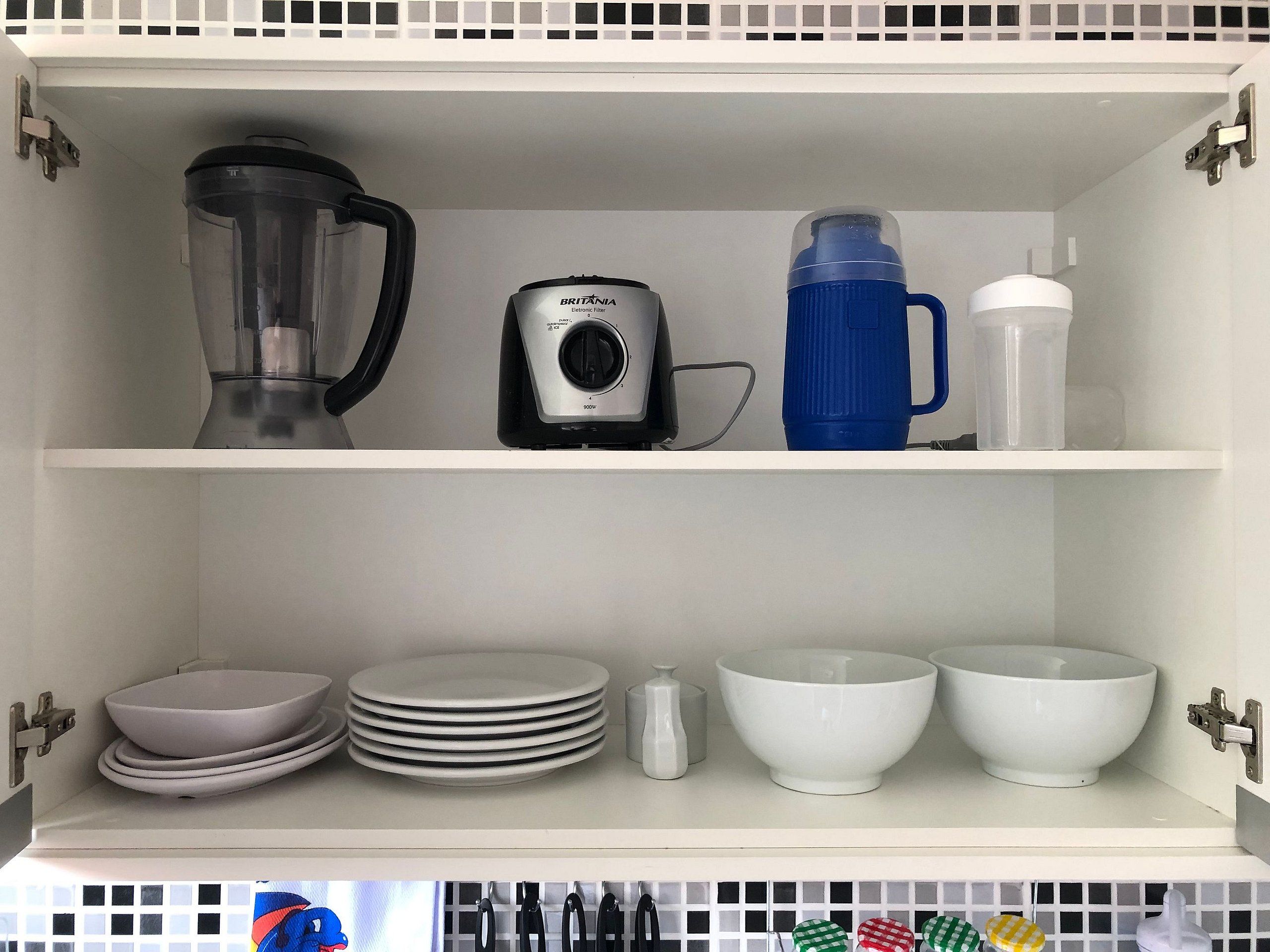 Una cocina con una licuadora, hervidor y botella de agua en el estante superior, y platos y tazones apilados en el inferior.