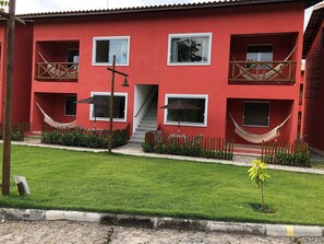 A red building with multiple windows, a staircase, and a fenced garden area with a hammock. - Apartamento na região de Guarajuba (Camaçari)