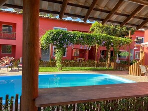 A swimming pool with a wooden deck, surrounded by a wooden fence, and a red building with windows and a balcony in the background.