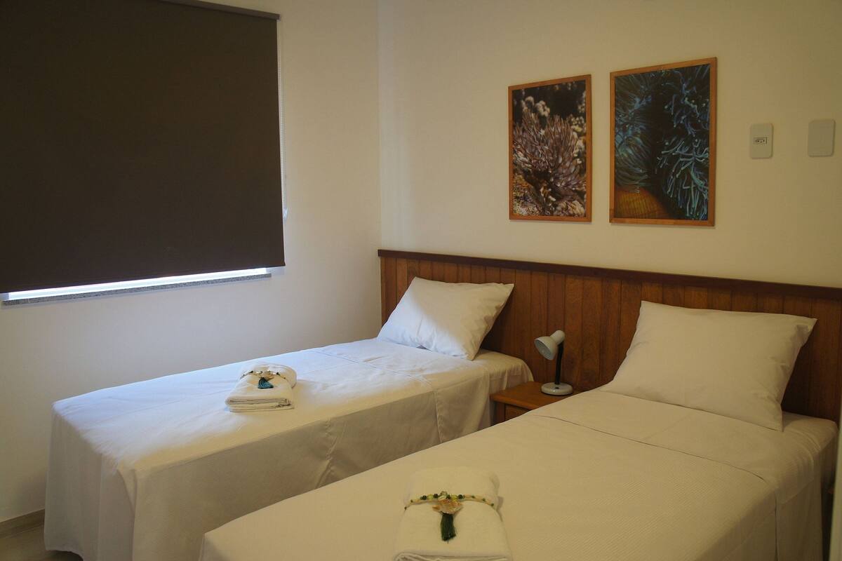 A hotel room with two single beds, a wooden headboard, a nightstand, a lamp, and two framed pictures on the wall.