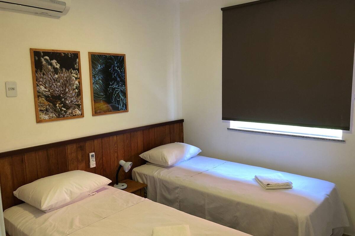A hotel room with two single beds, a wooden headboard, a nightstand with a lamp, and two framed pictures on the wall.
