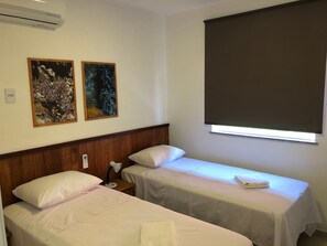 A hotel room with two single beds, a wooden headboard, a nightstand with a lamp, and two framed pictures on the wall. - Apartamento na região de Guarajuba (Camaçari)