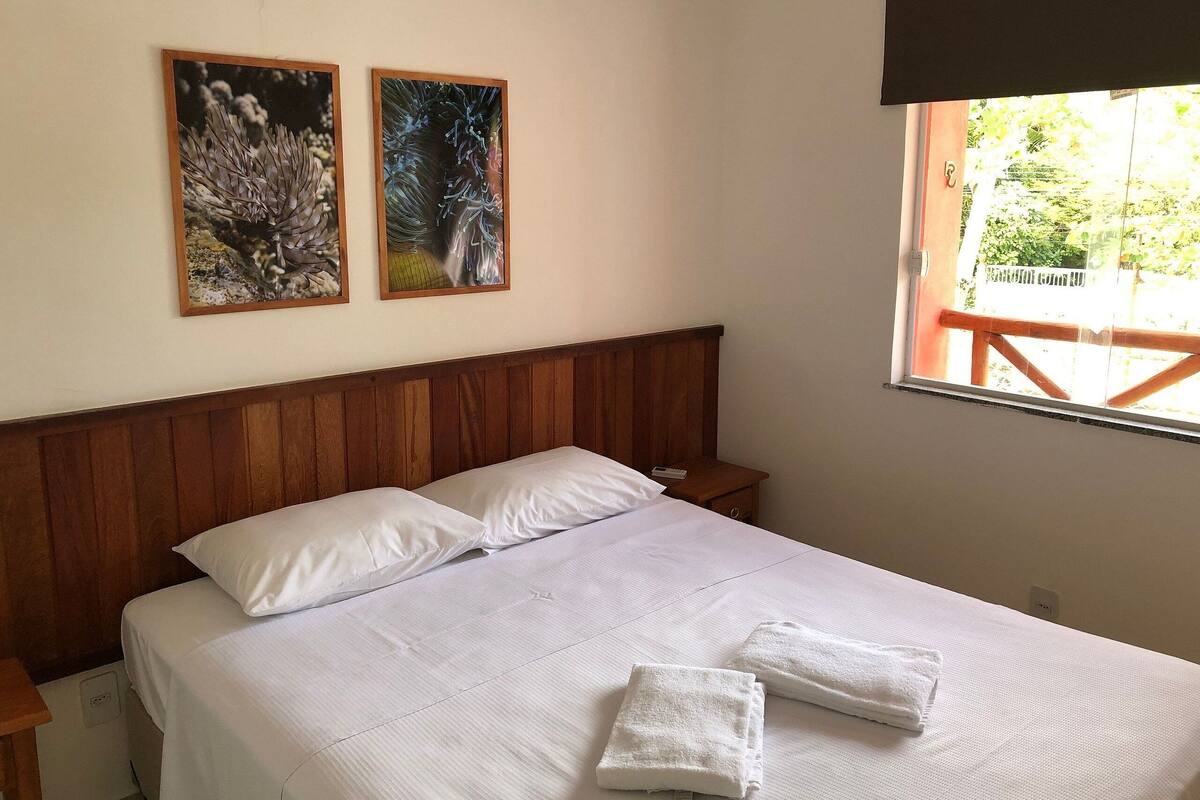 A neatly made bed with white linens and two pillows, a wooden headboard, and two framed artworks on the wall.