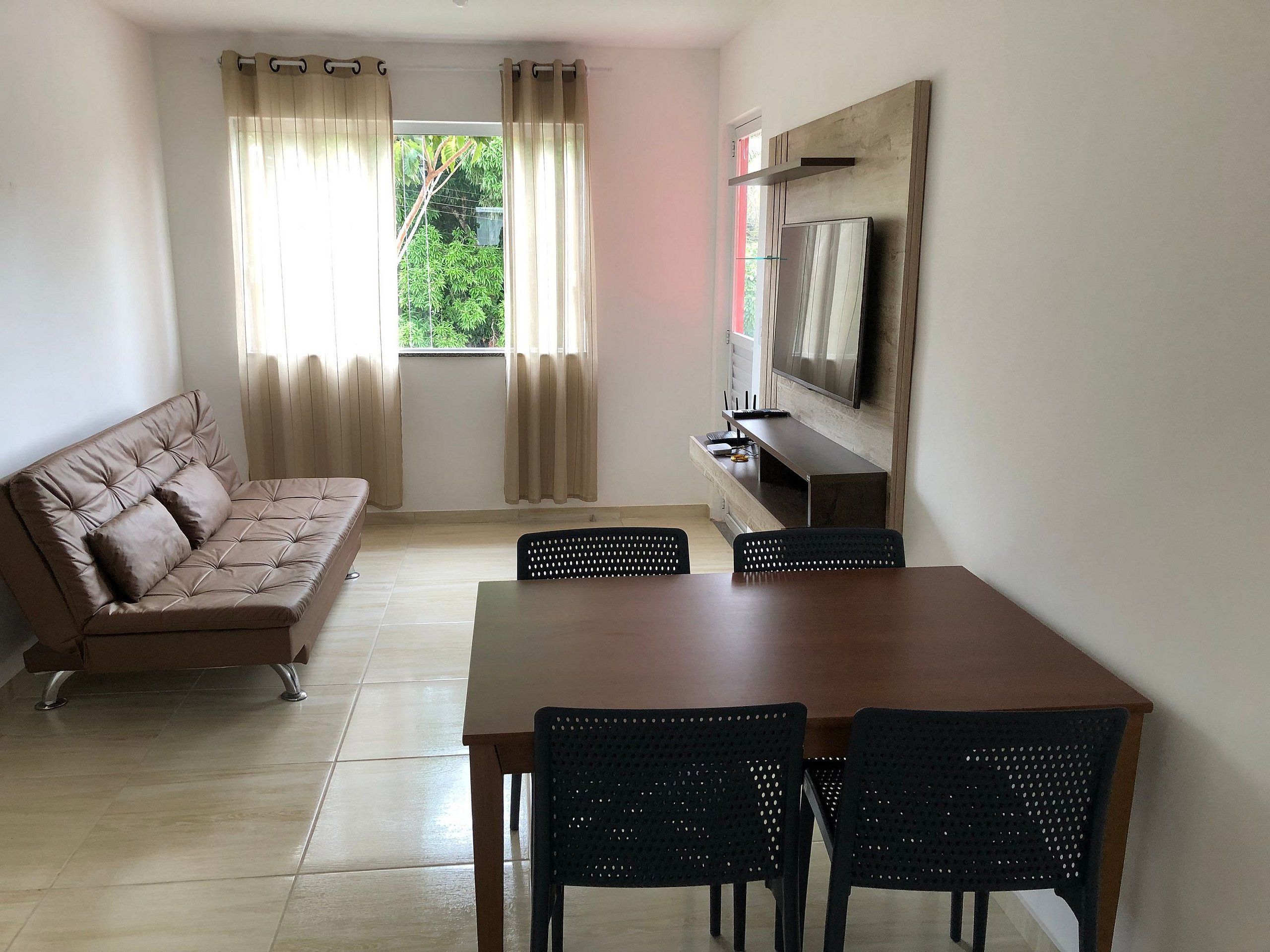 Una sala de estar con un sofá, un televisor instalado en la pared, una mesa de comedor con sillas y una ventana con cortinas.