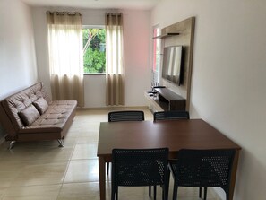 A living room with a sofa, a TV mounted on the wall, a dining table with chairs, and a window with curtains.