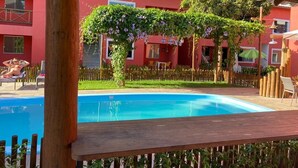 Una piscina con cubierta de madera, rodeada por una cerca de madera, y un edificio rojo con ventanas y un balcón al fondo.