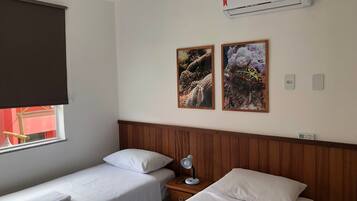 A hotel room with two single beds, a wooden headboard, a window with a blind, an air conditioner, two framed pictures on the wall, and a bedside table with a lamp.