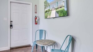 A room with a white door, a fire extinguisher, a round table with chairs, and a framed picture on the wall.
