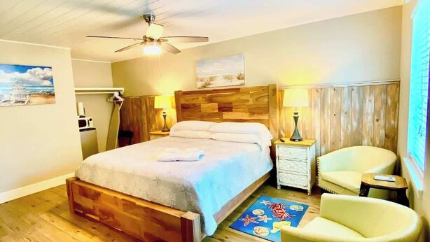 Quarto com cama de madeira, ventilador de teto, mesa de cabeceira, cadeira e uma pequena mesa.