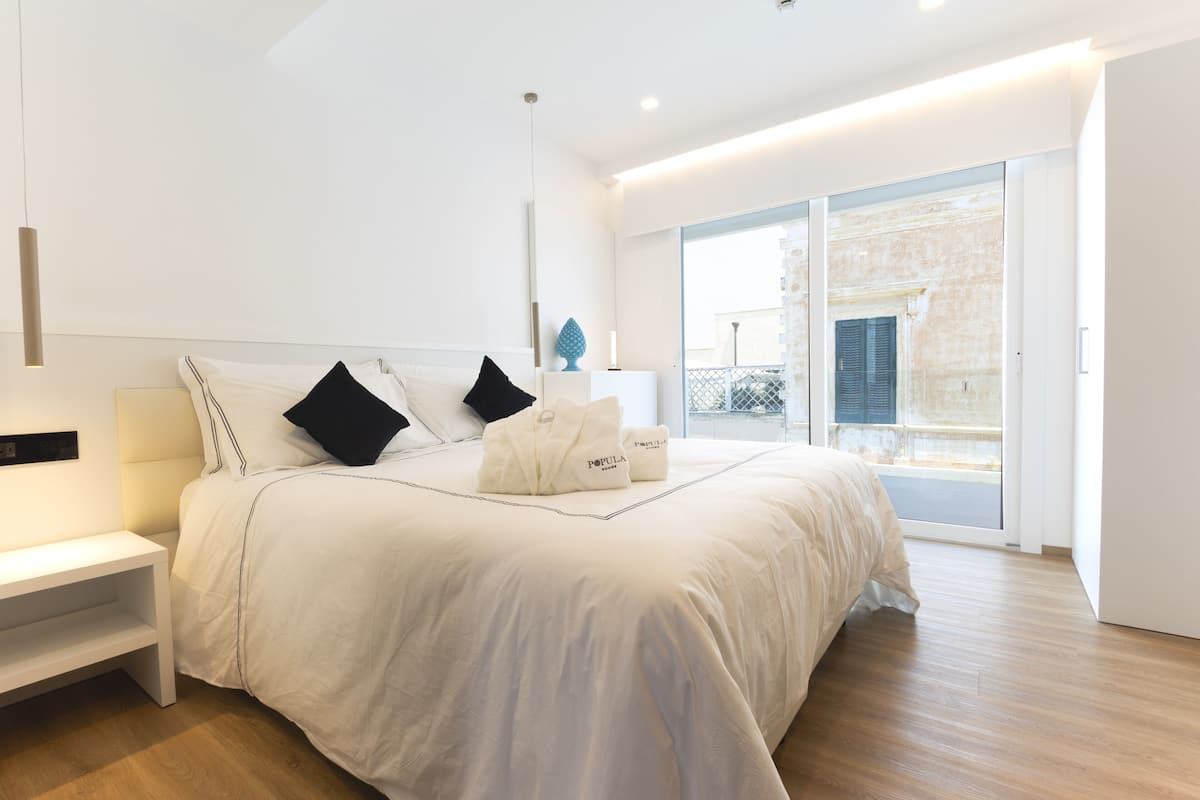 A modern bedroom with a large bed, white bedding, and black pillows. There is a bedside table and a lamp. The room has a wooden floor and a sliding glass door leading to an outdoor area.