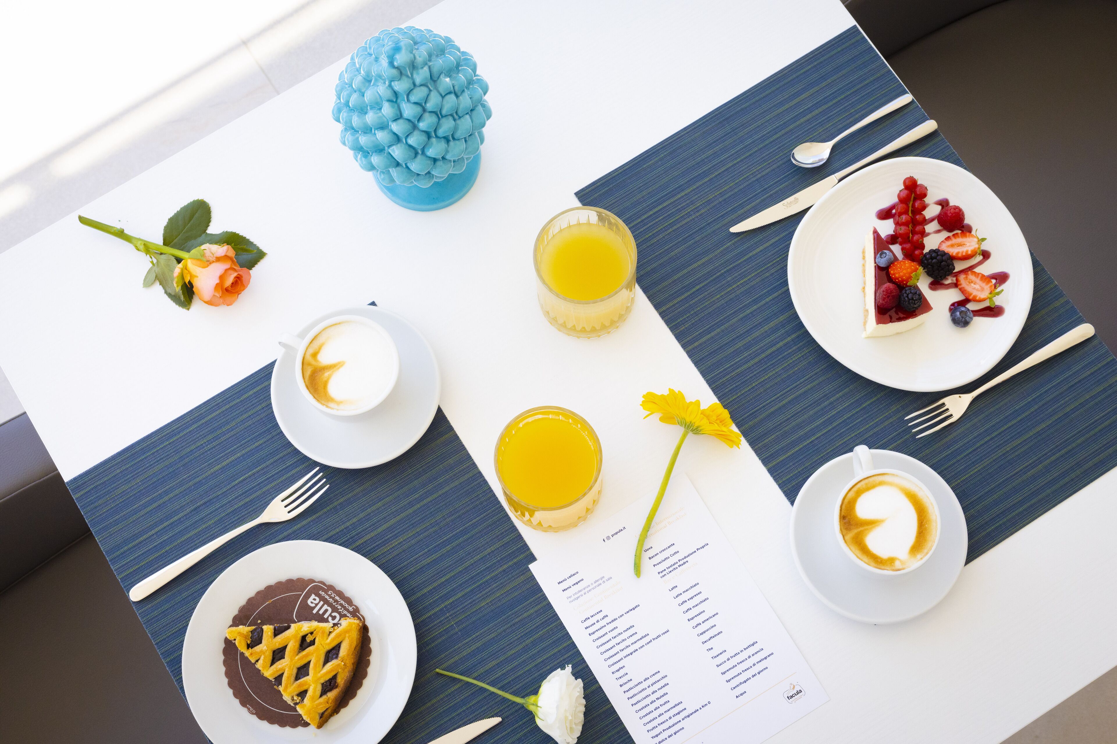 Un desayuno con un menú, un jarrón decorativo azul, una bebida amarilla y un plato de fruta y postre.