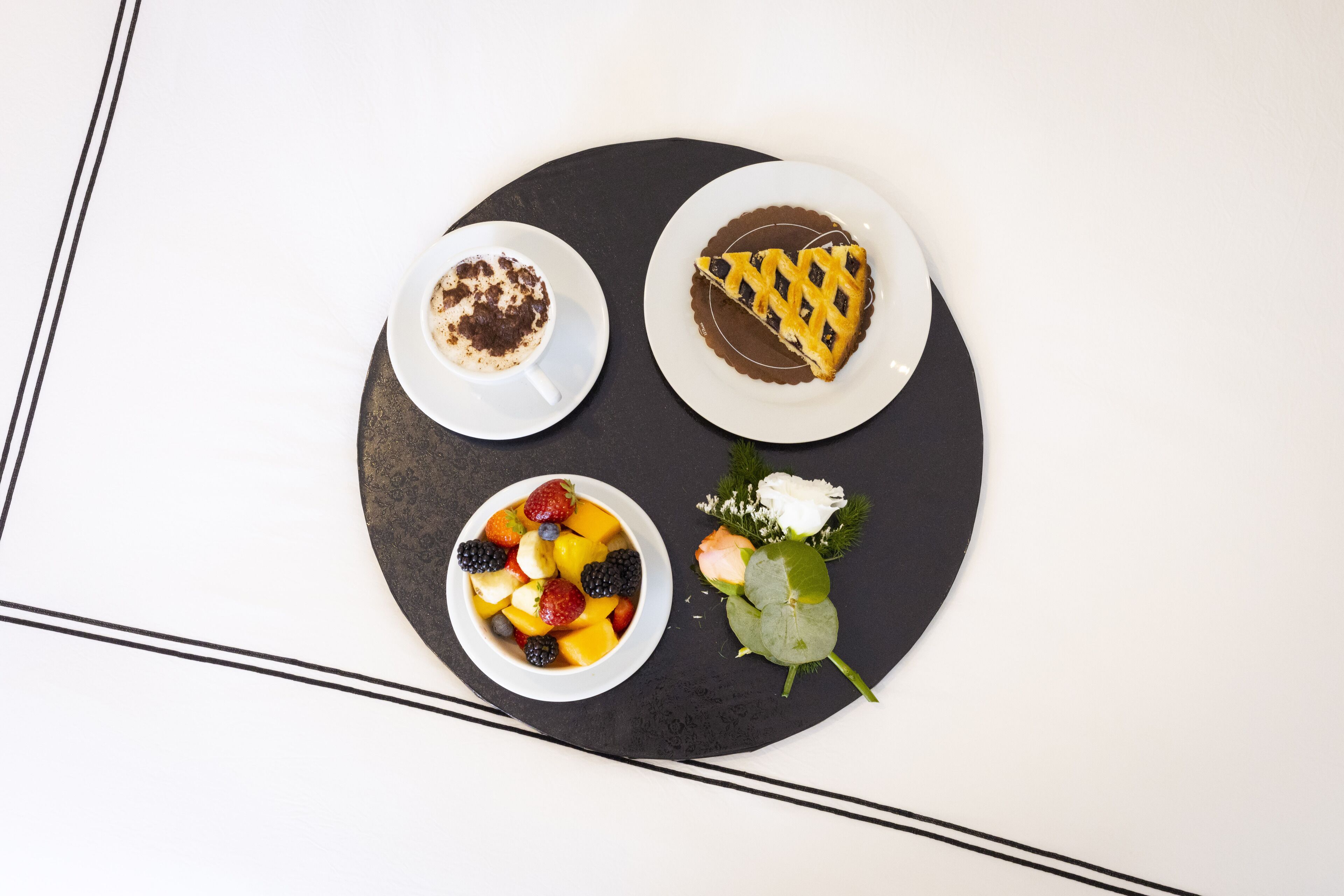 Une assiette à dessert avec une part de tarte, un bol de fruits et une tasse de café, le tout posé sur une table noire.