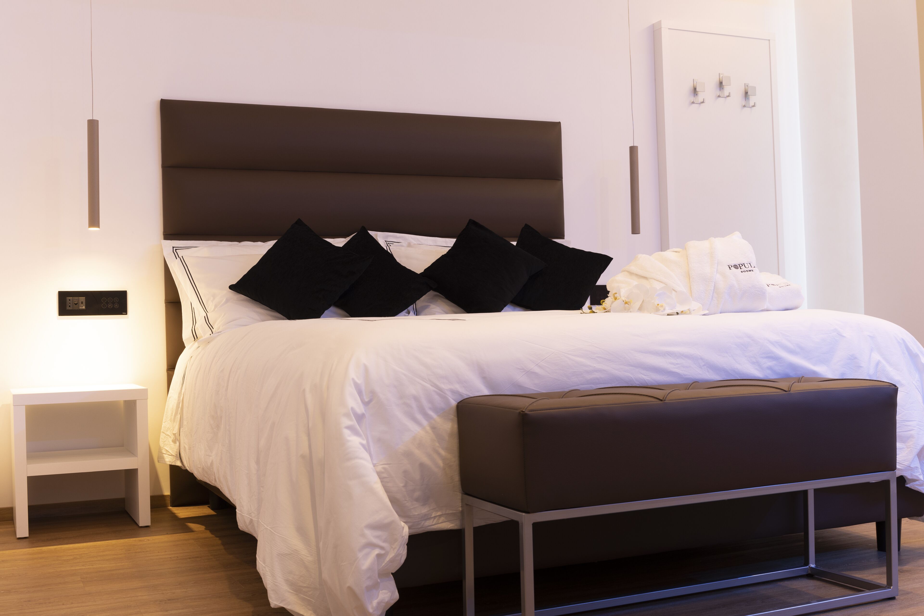 A neatly made bed with a white comforter and black pillows, a bedside table with a lamp, and a bench at the foot of the bed.