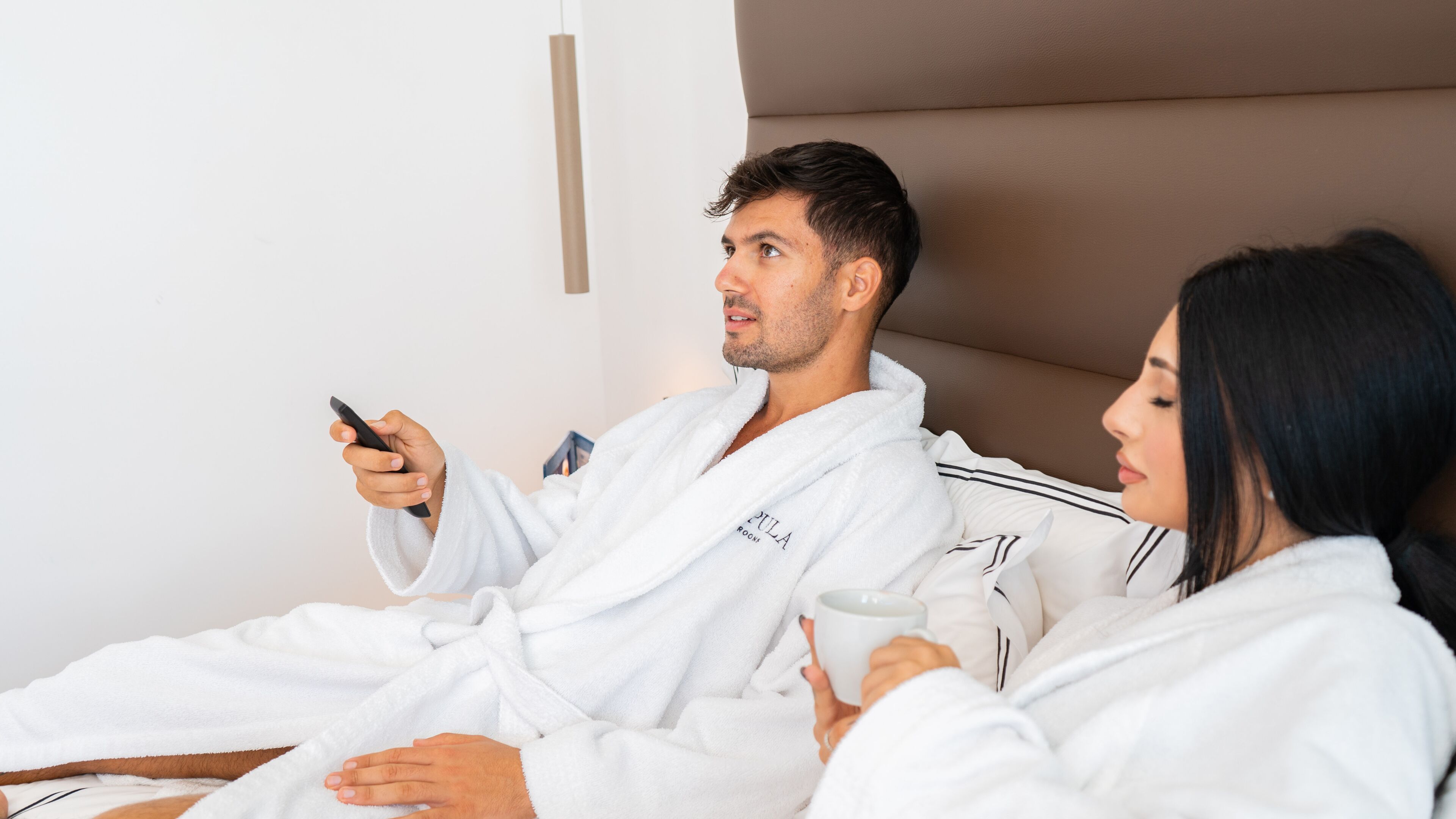 A couple in white bathrobes sitting on a bed in a room with a plain white wall and a brown headboard.