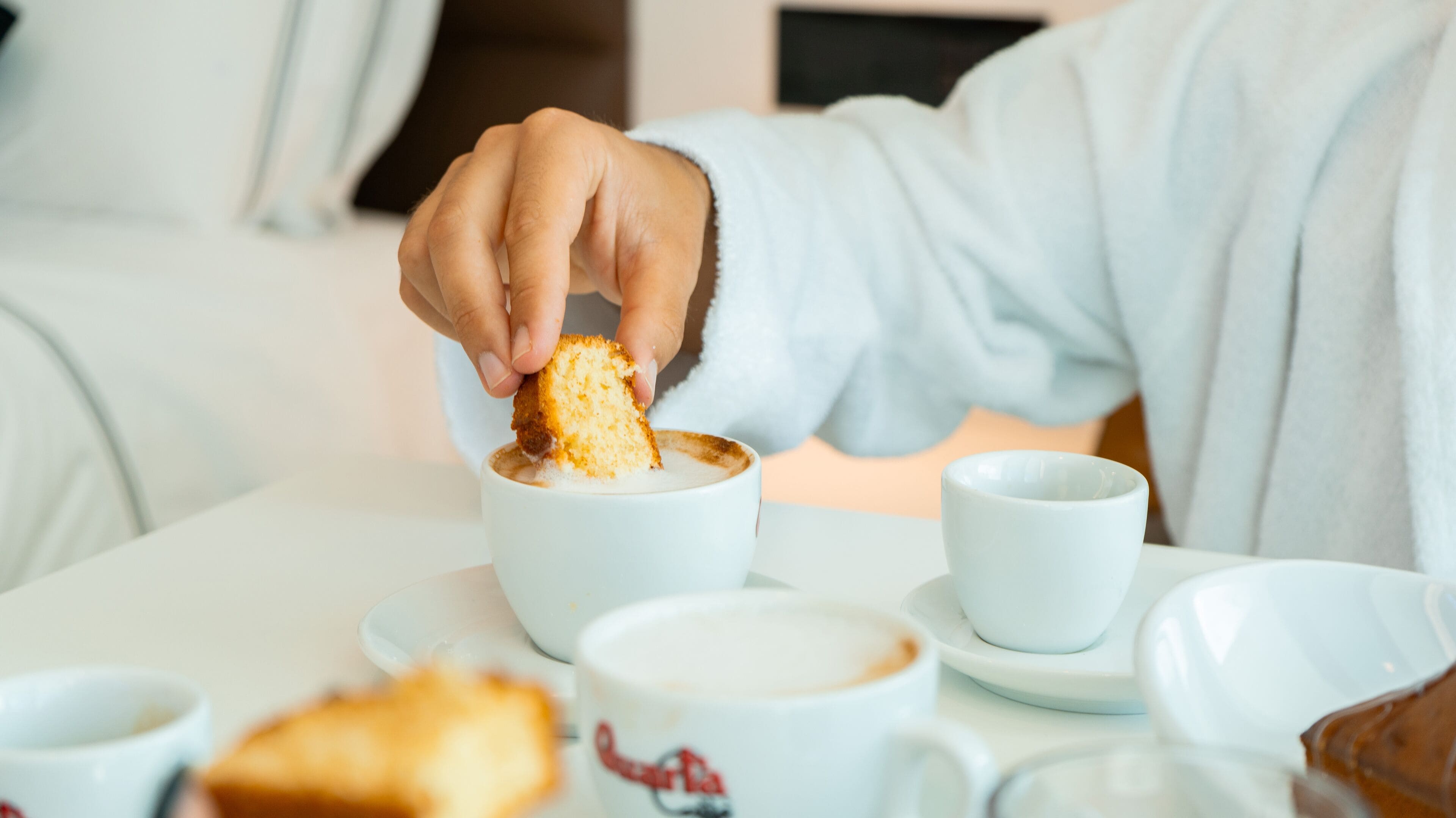 Una persona con bata blanca sentada a una mesa con una taza blanca, un platillo y un trozo de pan.