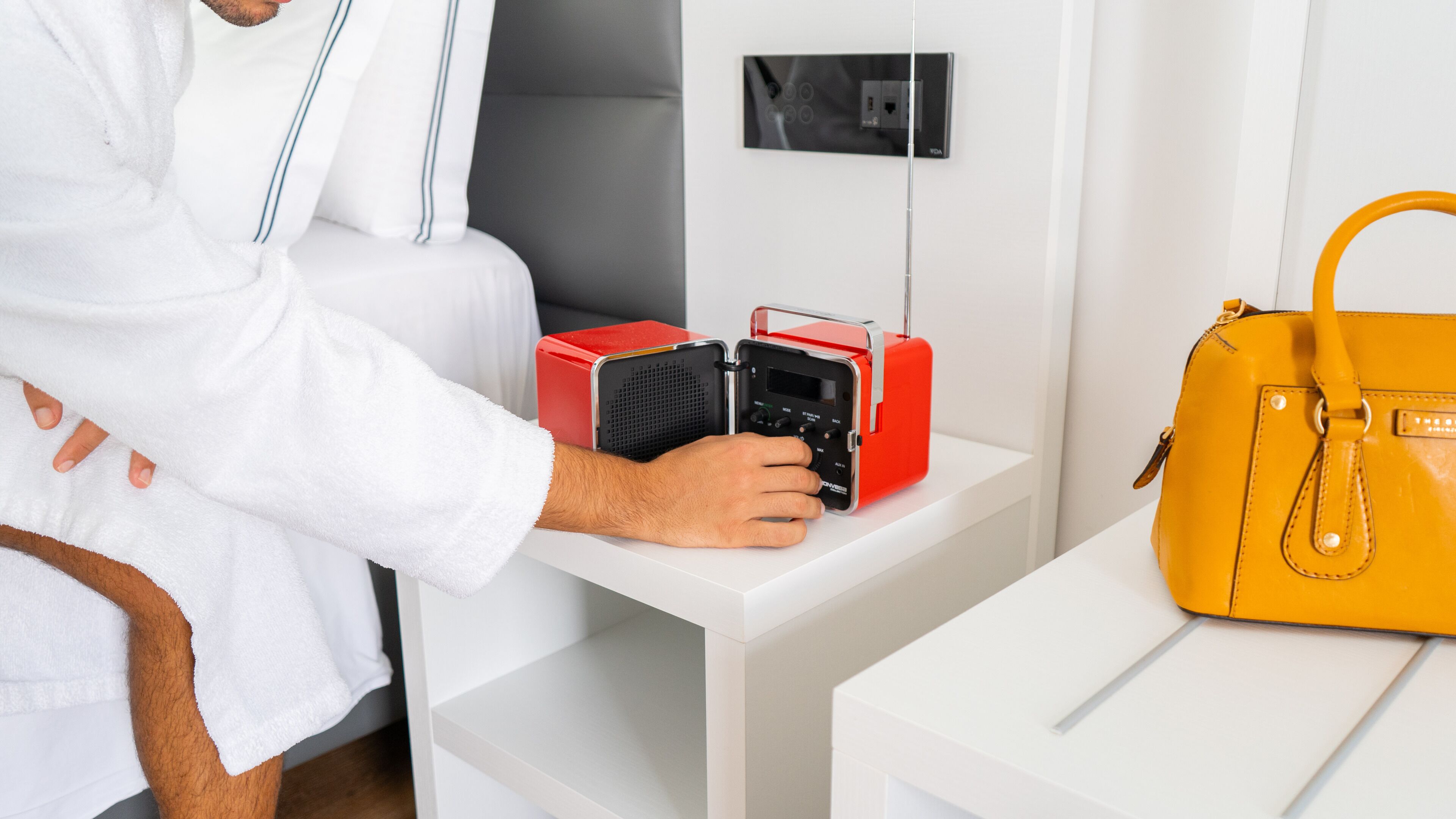 Eine Person in einem weißen Bademantel sitzt neben einem roten und schwarzen Radio auf einem weißen Nachttisch, mit einer gelben Handtasche auf einem weißen Beistelltisch.