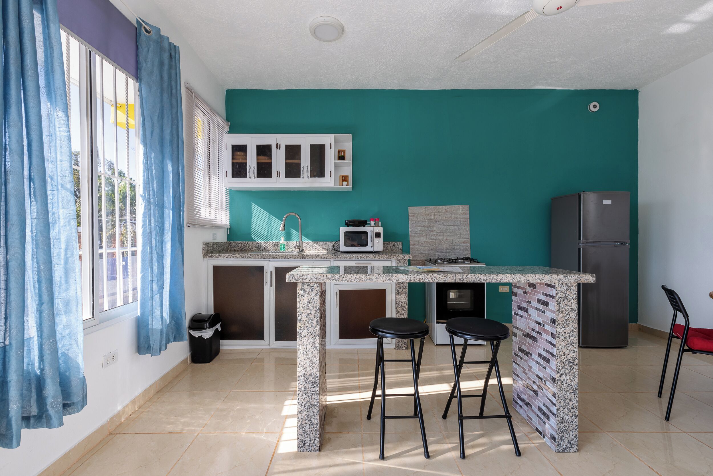 Una cocina compacta con pared verde azulada, un armario blanco, un microondas, un refrigerador y dos taburetes de bar.