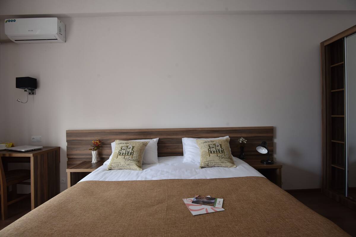 A neatly arranged hotel room with a wooden headboard, a bed with white linens, two decorative pillows, a bedside table with a lamp, a laptop, and a wall-mounted air conditioner.