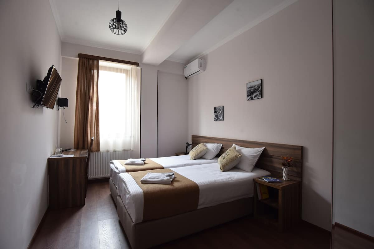 A hotel room with two beds, a desk, a chair, a radiator, an air conditioner, and a window with curtains.