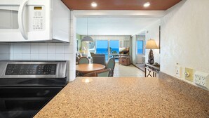 A kitchen with a microwave, stove, granite countertop, wooden cabinets, and a dining area with a table and chairs. - Captivating & Relaxing 5th Floor, Ocean Surrounds, A/C in Bedroom (Koloa)