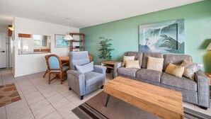A living room with a grey sofa, a blue armchair, a wooden coffee table, a small side table, and a painting on the wall. - Captivating & Relaxing 5th Floor, Ocean Surrounds, A/C in Bedroom (Koloa)