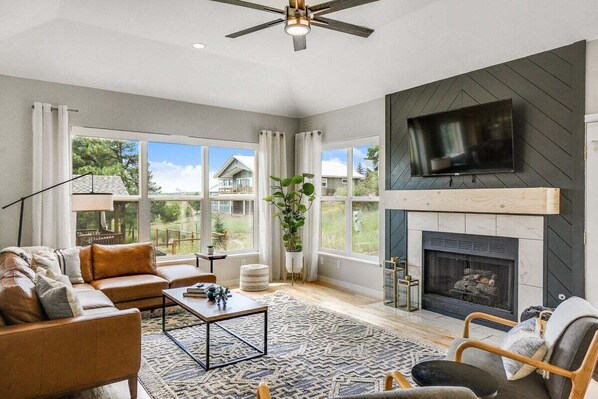 A spacious living room with a fireplace, large windows, and a flat-screen TV mounted on the wall. - Large Home, Hot Tub, Close to Lakes and Shopping (Pagosa Springs)