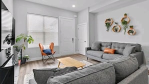 A modern living room with a grey sofa, a wooden coffee table, and a chair. There are potted plants on the wall and a TV on the left.