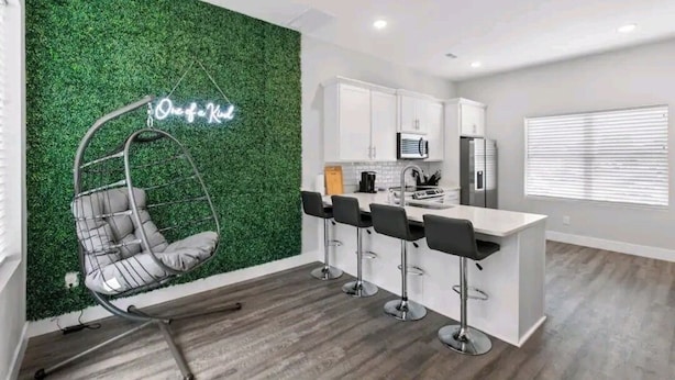 A modern kitchen with white cabinets, a bar area with stools, and a hanging chair with a neon sign.