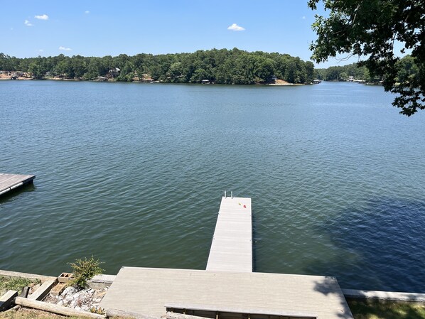 A calm lake with a wooden dock extending into the water, surrounded by trees and a distant shoreline with buildings.