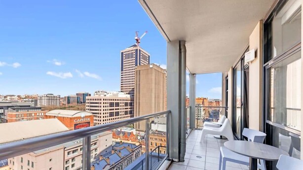A balcony with a city view, featuring a high-rise building with a crane, a red building with 'CAPITOL' signage, and a tiled floor with white chairs and tables.