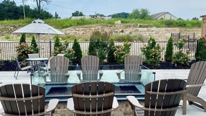Een terras buiten met Adirondack-stoelen, een tafel met glazen blad en uitzicht op een zwembad en het landschap.