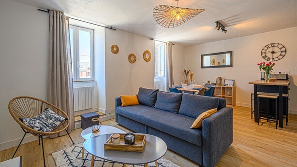 Una sala moderna con un sofá gris, una mesa de centro redonda y una silla de mimbre. Hay una zona de comedor con mesa y sillas, un reloj en la pared y una ventana con cortinas.