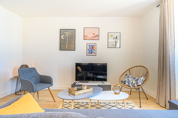 A modern living room with a flat-screen TV, a grey armchair, a wicker chair, and a coffee table.