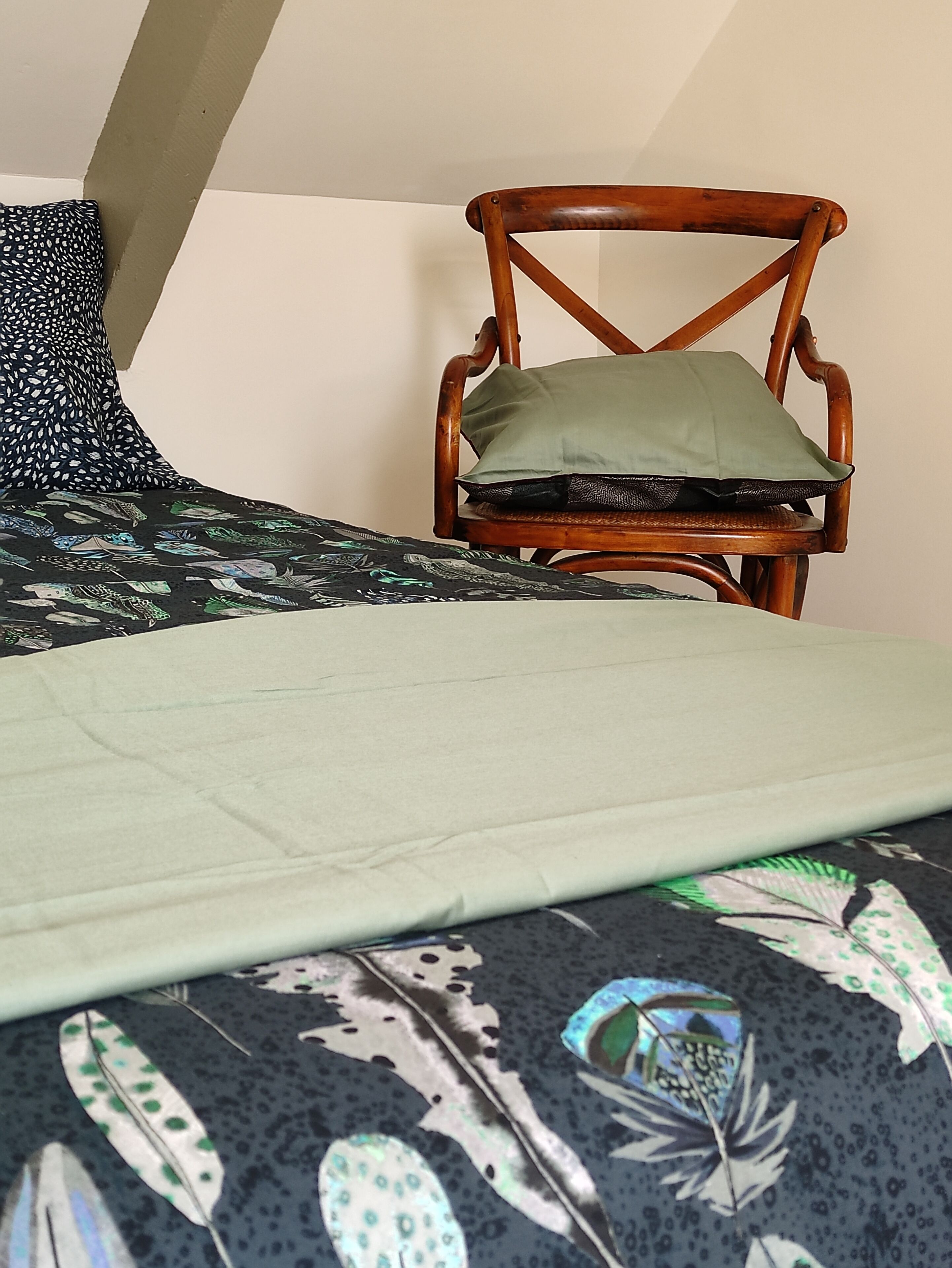 Ein Zimmer mit einem Bett, einem Holzstuhl mit Kissen und einer gemusterten Tagesdecke.