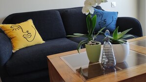 A living room with a dark blue sofa, a yellow pillow with a face design, a blue pillow with a face design, a green plant, a white orchid, and a wooden coffee table.