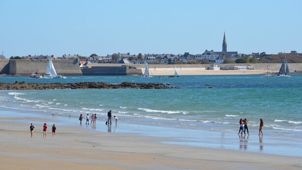 A beach with people walking along the shore, a lighthouse in the background, and sailboats on the water.