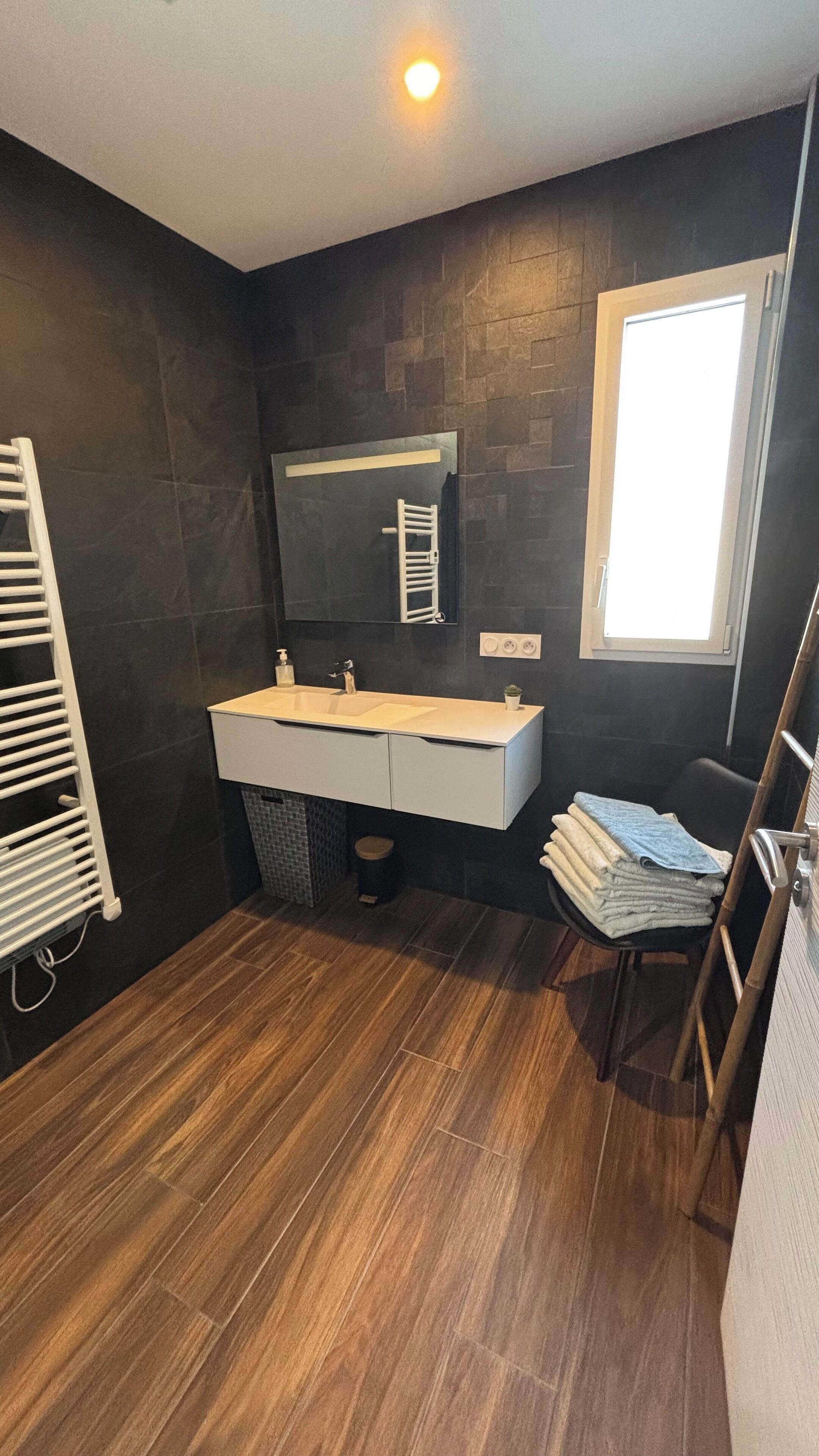 A modern bathroom with a dark tiled wall, a white vanity, a large mirror, and a wooden floor.